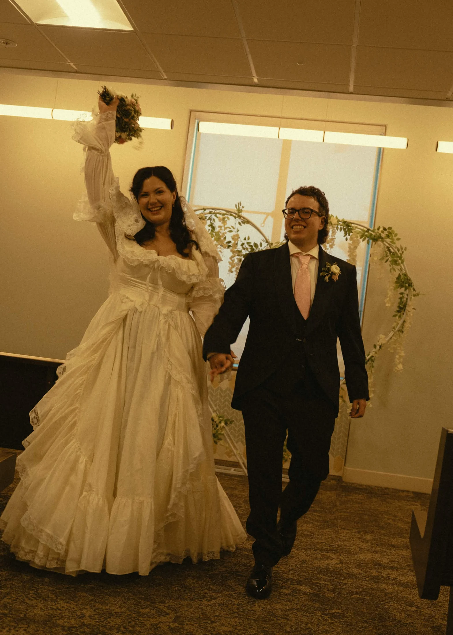 Samantha raising her bouquet as she and Stephen walk hand in hand after getting married